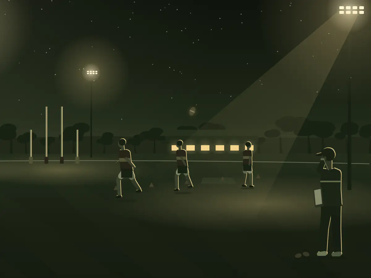 A footy coach blows the whistle on a kicking drill under the lights of a country oval.